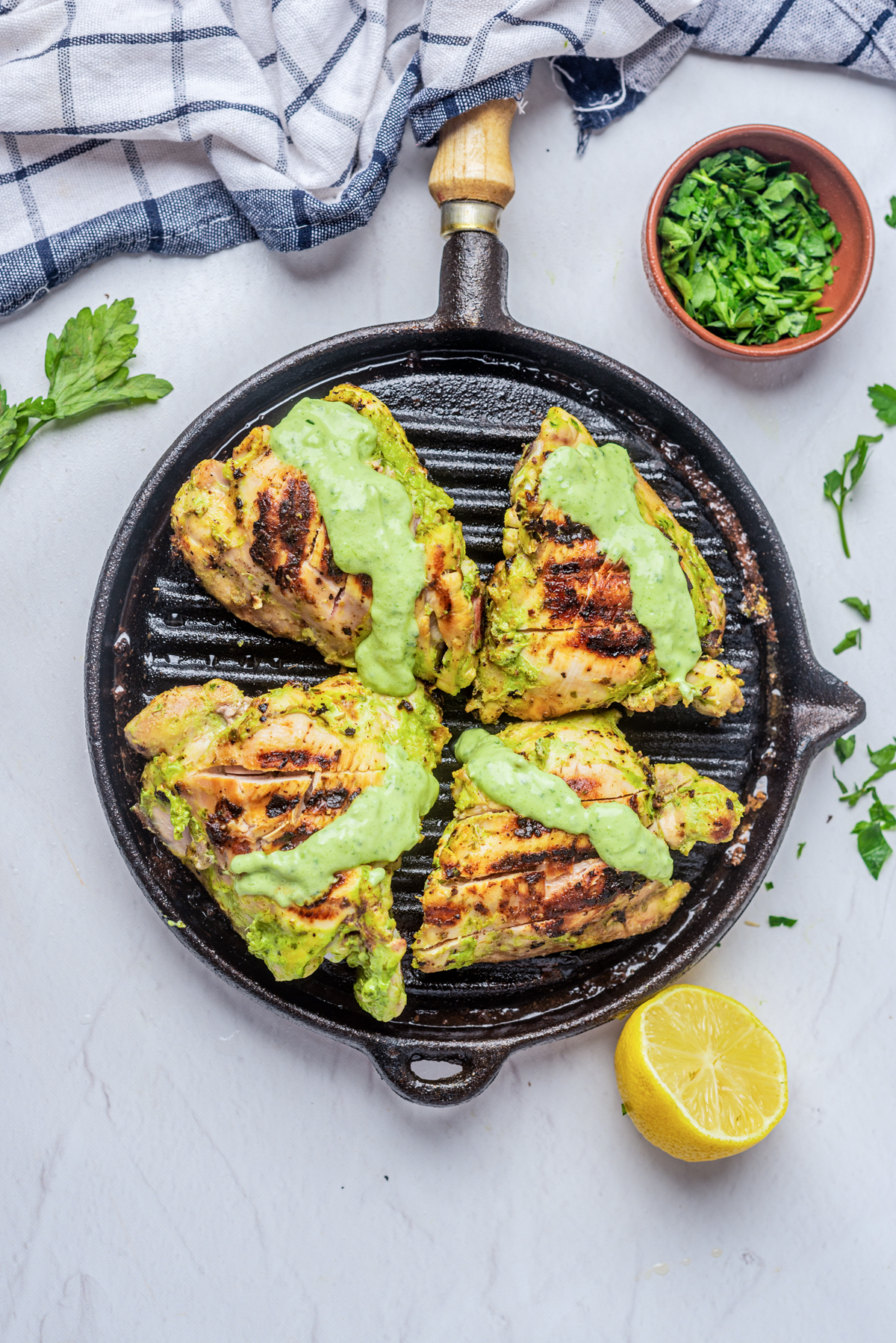 4 green goddess chicken thighs on a grill pan with dressing on top, on a white background, half lemon bottom right, green herbs in a small wooden bowl top right, blue and white striped linen along the top