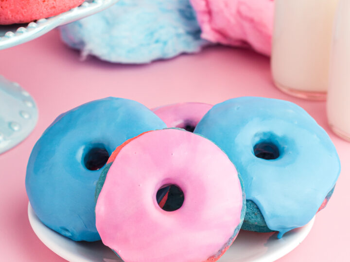 2 blue and 1 pink (in center) donuts on a white plate. blue and pink cotton candy in the background middle, 2 milk jugs on the right, cake stand on the left showing a bit of donuts