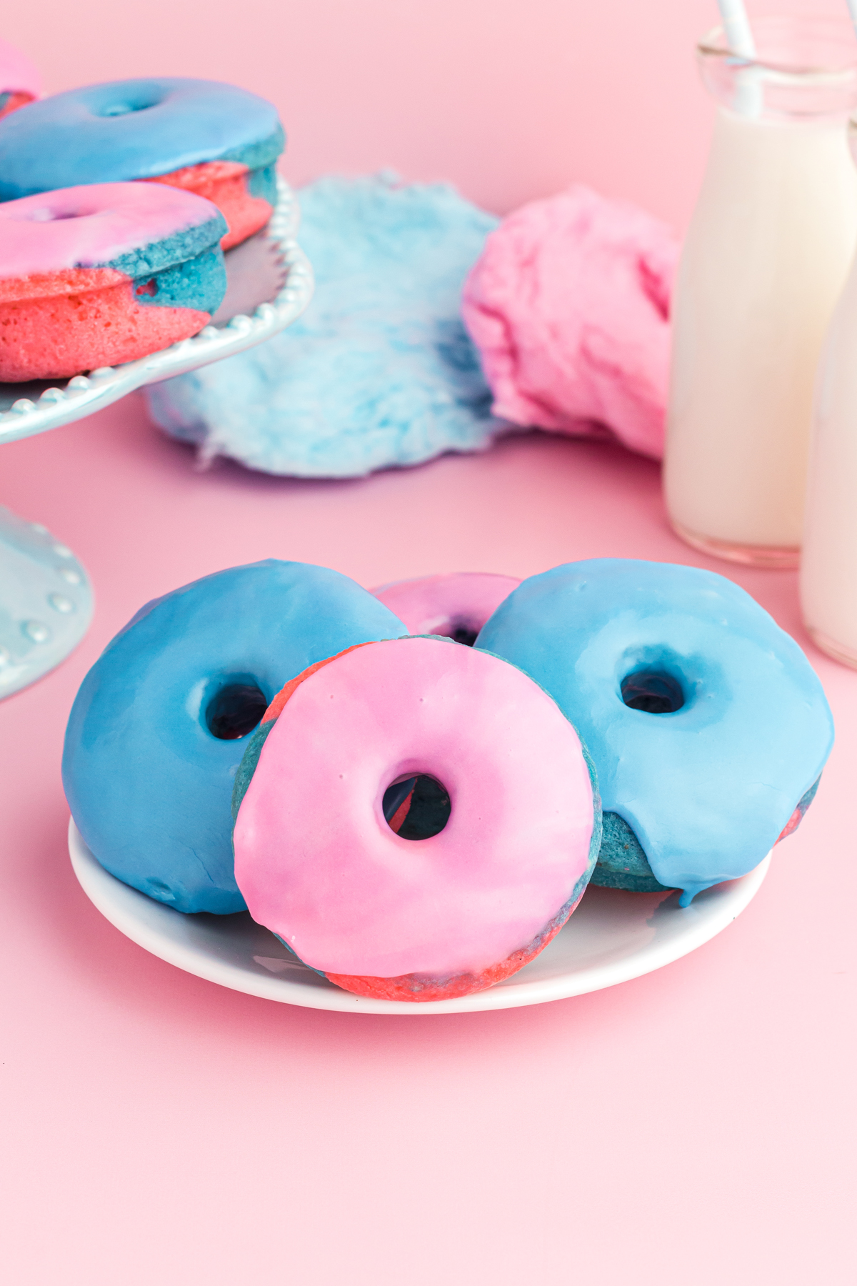 Baked Donuts Recipe - 2 blue and 1 pink (in center) donuts on a white plate. blue and pink cotton candy in the background middle, 2 milk jugs on the right, cake stand on the left showing a bit of donuts