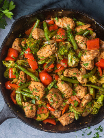 Chicken breast bites, red bell peppers, zucchini, green beans and broccoli cooked in a cast iron pan with chopped parsley on top and in the top left corner