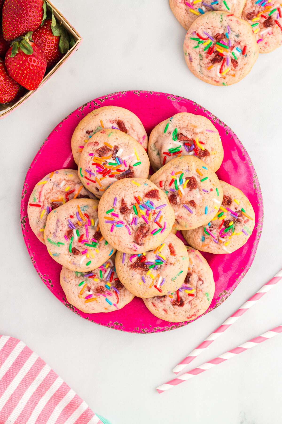 stack of strawberry milkshake cookies on a hot pink plate. 6 cookies on 1st layer, 4 on next layer and one top layer. top left corner strawberries peeking out, top right a cookie, bottom right red & white striped straws, bottom left red and white striped cloth peeking out.